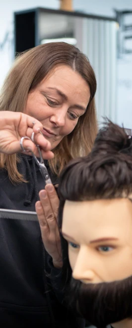 A female barbering student in a black hoodie carefully cutting the hair on a mannequin head inside a training facility. A female barbering student in a black hoodie carefully cutting the hair on a mannequin head inside a training facility.
